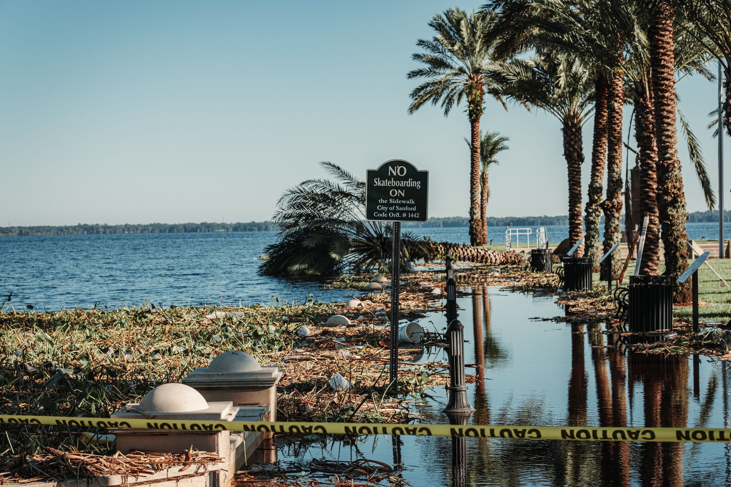 A flooded pathway beside a lake in Sanford, Florida, closed off with caution tape, palm trees standing in the water.