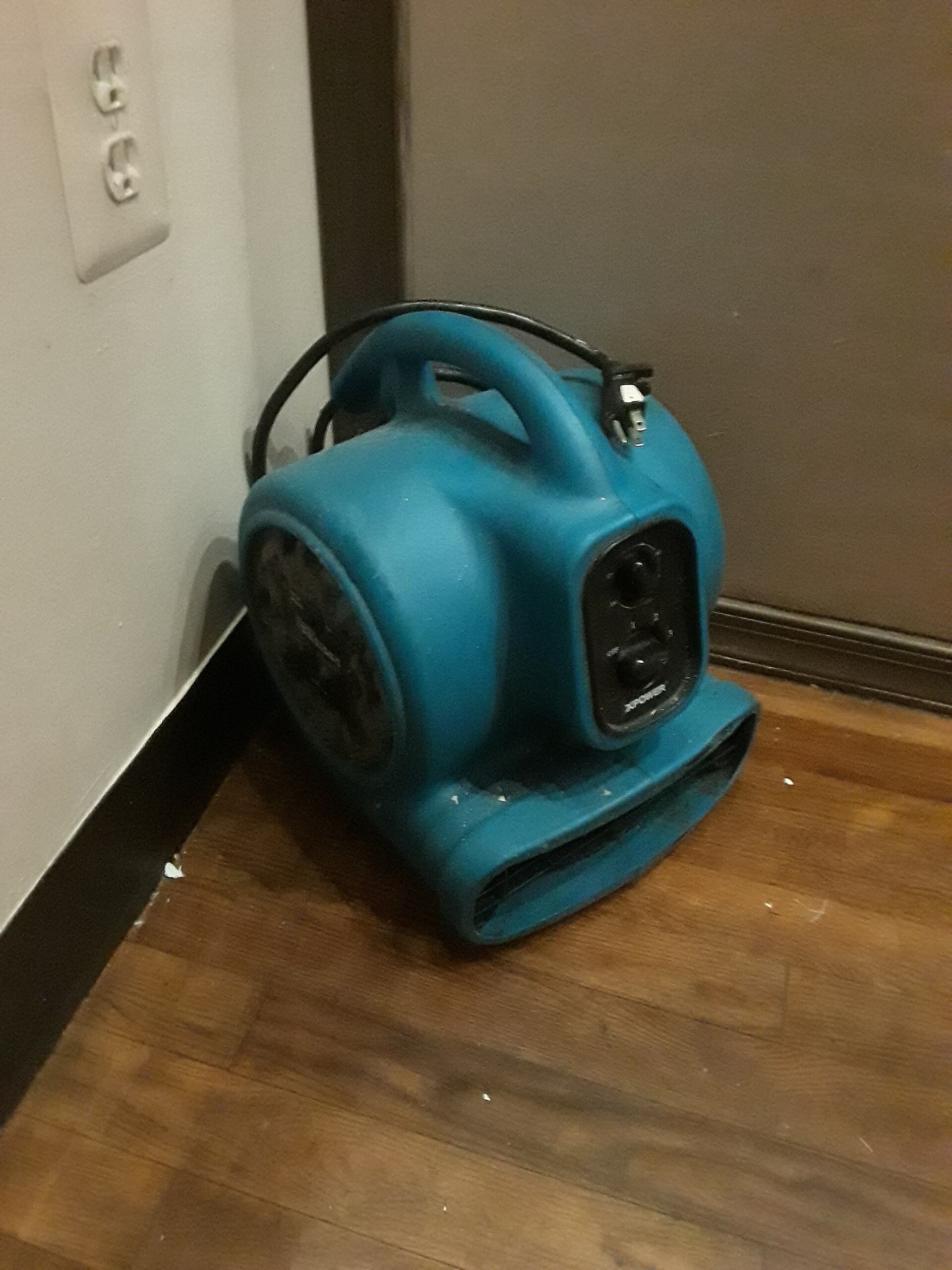 A blue XPOWER air mover angled into the base of a wall on a wood floor, plugged into a nearby outlet, with plaster dust on the boards.