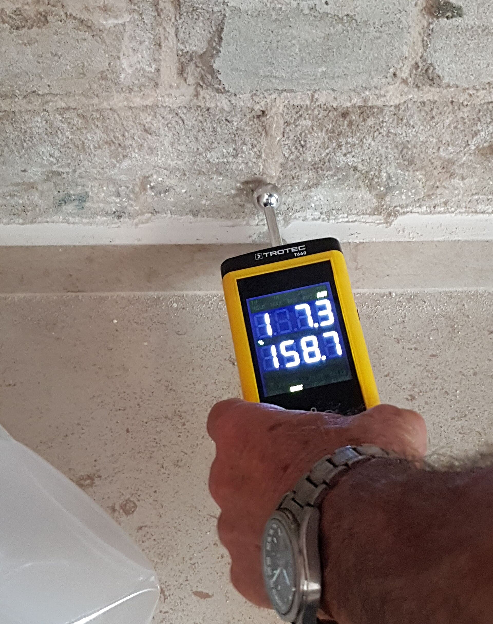 A hand holding a yellow Trotec T660 moisture meter against a rough masonry wall, its screen showing two numeric readings.